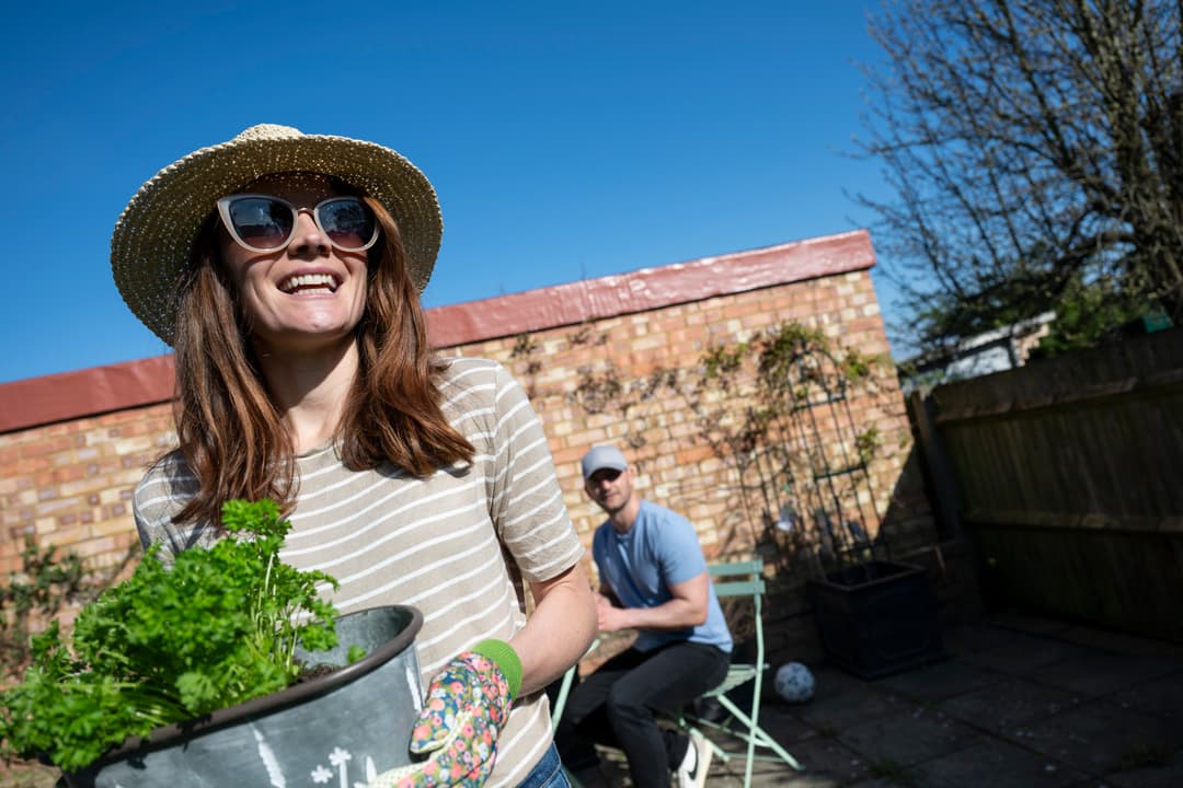 Woman wearing a hat and gardening gloves and holding a flower pot.
