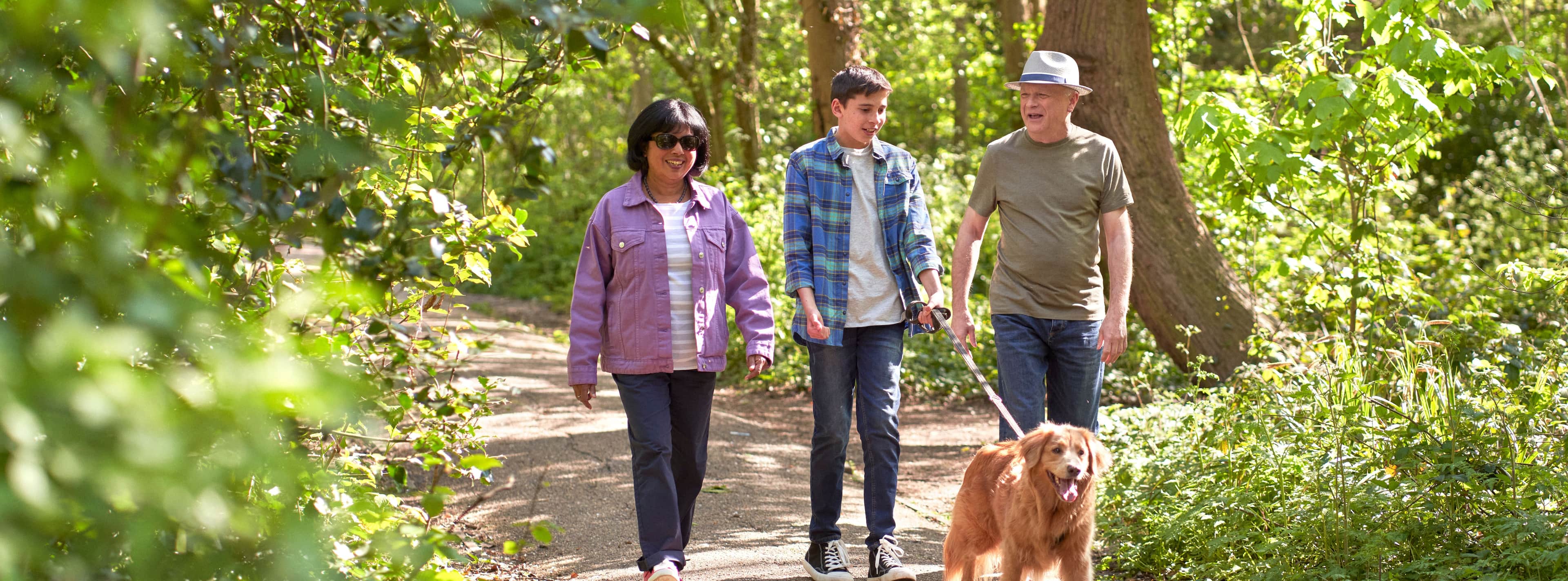 3 people walking outside with a dog in an area with shade.